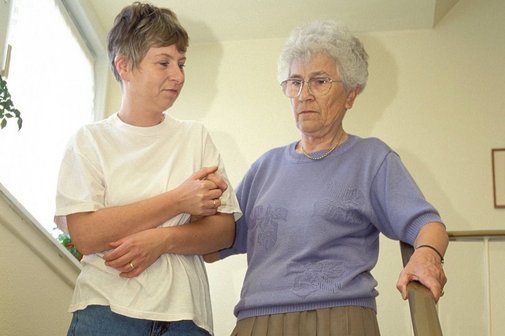 Eine jüngere Frau hilft einer Seniorin, eine Treppe hinabzugehen. Sie hält die ältere Frau am Arm eingehakt fest.