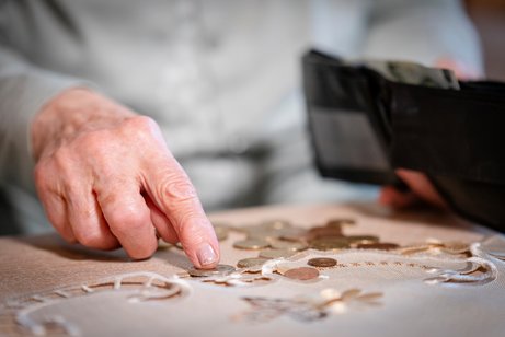 Eine Person z&auml;hlt einzelne M&uuml;nzen auf einem Tisch; eine Hand legt eine M&uuml;nze ab, im Hintergrund ist ein ge&ouml;ffnetes, nahezu leeres Portemonnaie zu sehen &ndash; Symbolbild f&uuml;r Armut.