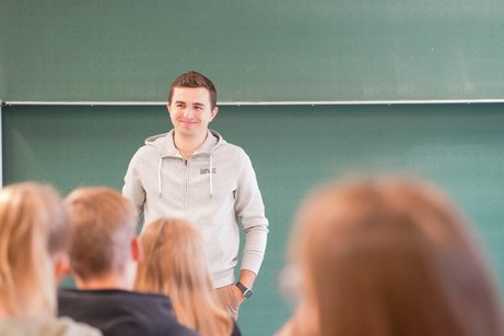 Ricardo Scheuerer steht vor einer grünen Tafel, blickt lächelnd in eine Gruppe von Zuhörenden bei einem Vortrag an der Uni Paderborn.