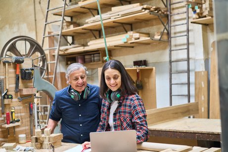 Ein &auml;lterer Mann und eine Frau in einer Schreinerei, sie beugen sich gemeinsam &uuml;ber ein Notebook, im Hintergrund Holz, Werkzeuge und Maschinen