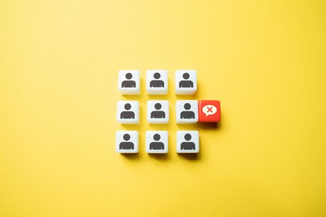 Symbolbild für Diskriminierung am Arbeitsplatz: Neun Würfel mit einem Personen-Icon sind in einem 3x3-Raster auf gelbem Hintergrund. Neben einer der Figürchen ist eine durchgestrichene Sprechblase.