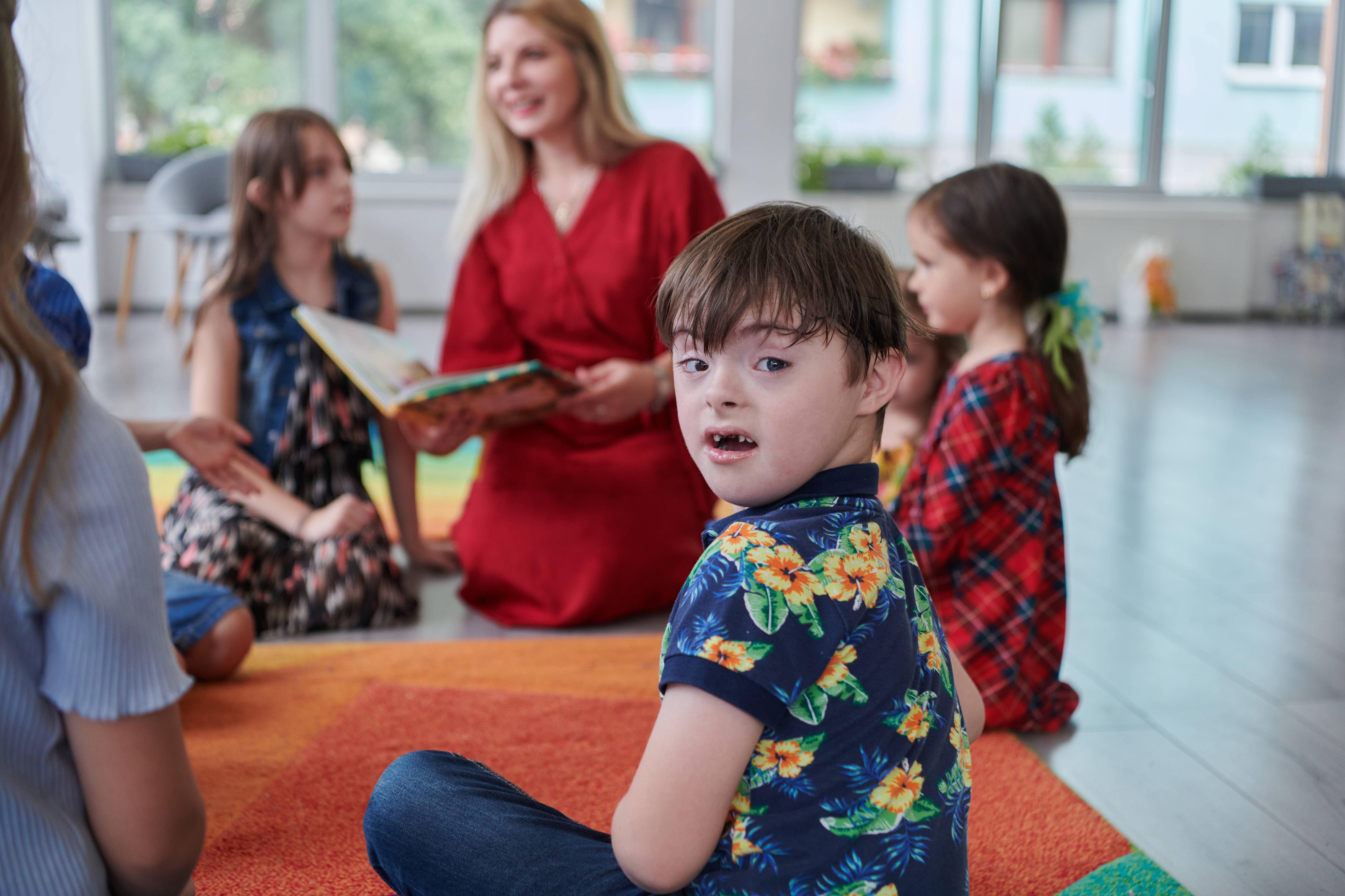 Inklusives Lernen Das Bild zeigt eine Schulklasse mit mehreren Kindern und einer Lehrerin, die aus einem Buch vorliest. Im Vordergrund sieht man einen Jungen mit Down-Syndrom, der sich umdreht und direkt in die Kamera schaut.
