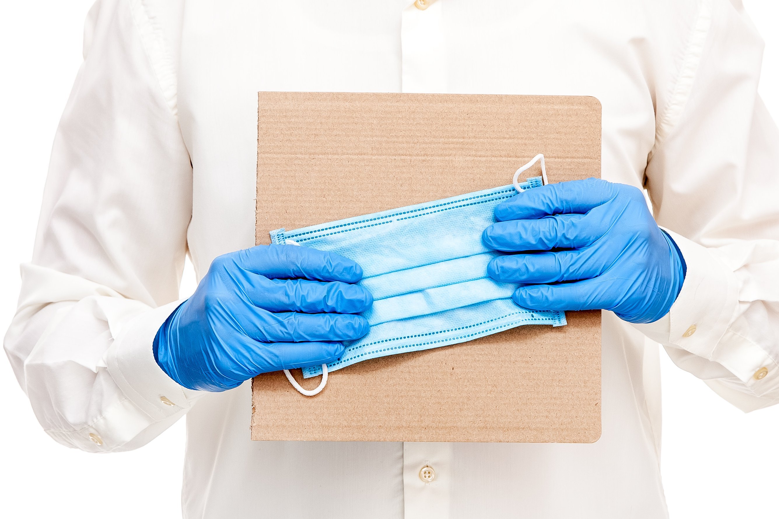 Pflegehilfsmittel Ein Mann im weißen Hemd trägt blaue Einweg-Handschuhe. Vor der Brust hält er einen schlichten Pappkarton und eine medizinische Schutzmaske.
