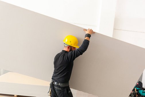 Ein Mann mit gelbem Bauarbeiterhelm trägt eine große Rigips-Platte auf einer Baustelle.
