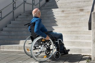 Ein älterer Mann im Rollstuhl steht ratlos am Fuß einer hohen Treppe, er schaut die Stufen hinauf. 