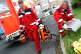 Zwei Rettungsassistenten der Feuerwehr im Rettungseinsatz, beide Männer tragen rote Berufskleidung und Ausrüstung, sie sind nur verschwommen zu erkennen.