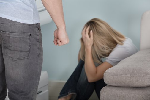 Hilfe für Opfer von Gewalt Gewalt in der Partnerschaft: Eine Frau sitzt neben einem Sofa auf dem Boden, schützt den Kopf mit den Händen. Im Vordergrund steht ein Mann mit geballter Faust.