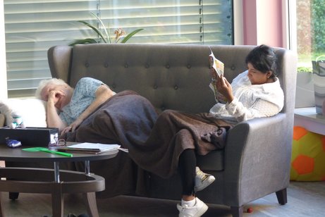 In der Demenz-WG mit dem Namen "Gammel-Oase" schläft eine ältere Dame auf einer Couch. Es ist mitten am Tag. Eine graue Decke ist über ihre Beine gelegt. Daneben sitzt eine Pflegeperson, die sich in eine Zeitschrift vertieft hat.