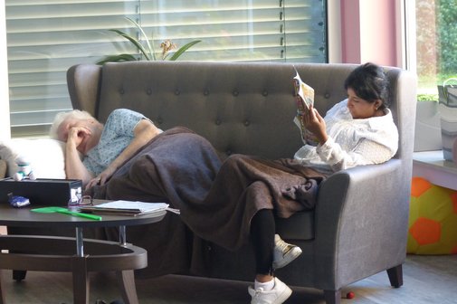 In der Demenz-WG mit dem Namen "Gammel-Oase" schläft eine ältere Dame auf einer Couch. Es ist mitten am Tag. Eine graue Decke ist über ihre Beine gelegt. Daneben sitzt eine Pflegeperson, die sich in eine Zeitschrift vertieft hat.