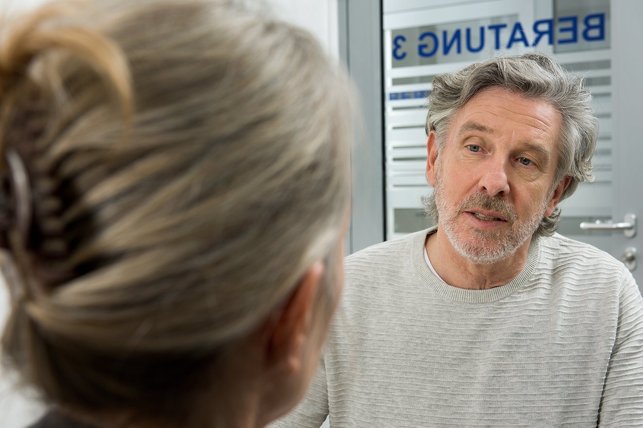 VdK-Beratung Ein Mann spricht mit einer Frau. Im Hintergrund erkennt man auf einer Glas-Trennscheibe das Wort "Beratung".