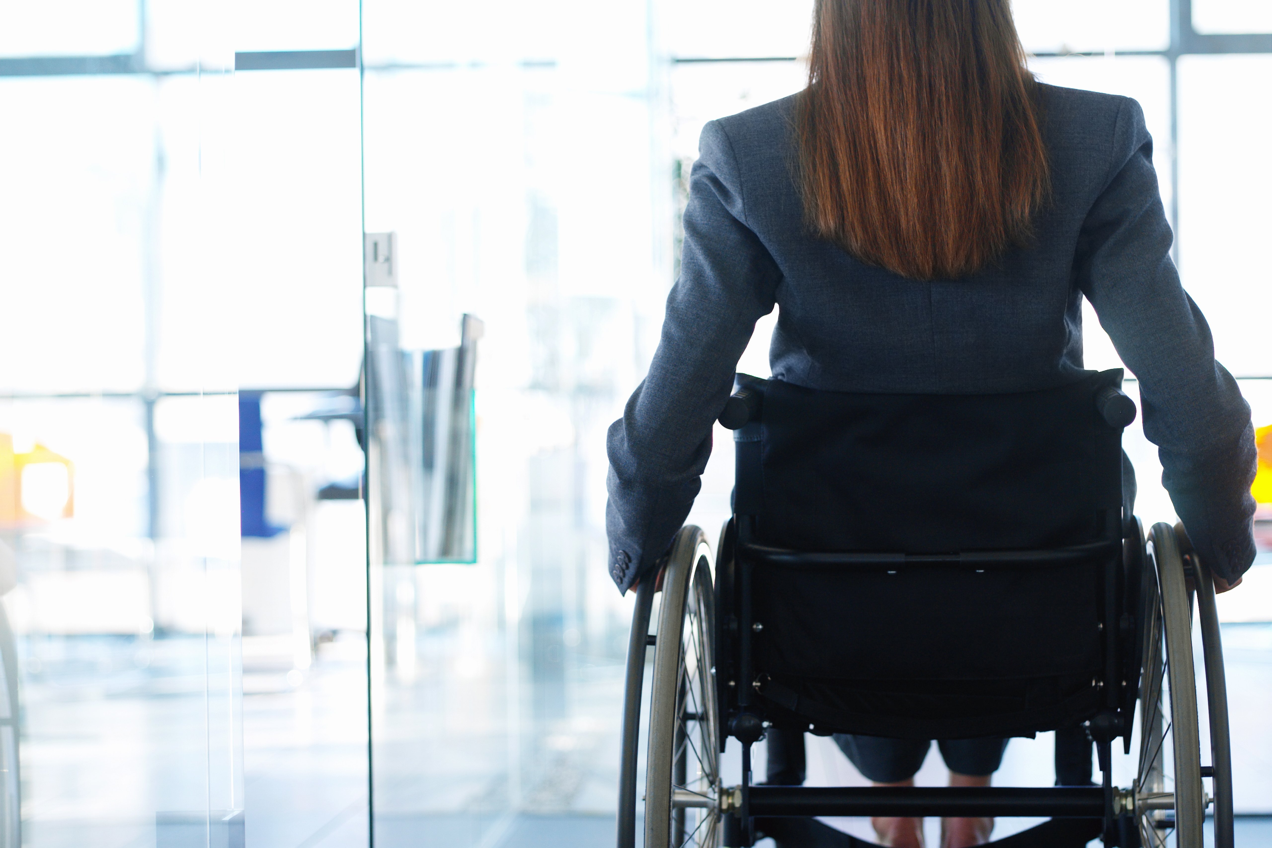 Rückenansicht einer Frau im Rollstuhl, sie befindet sich in einem Bürogebäude