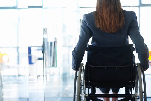 Rückenansicht einer Frau im Rollstuhl, sie befindet sich in einem Bürogebäude