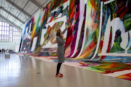 Stefanie Reimers steht in der Ausstellungshalle, betrachtet ein großformatiges Bild und spricht ins Telefon.