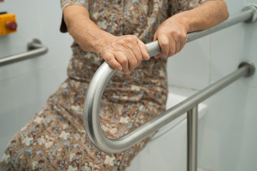 Eine &auml;ltere Frau stzt auf einem geschlossenen Toilettendeckel, sie h&auml;lt sich mit beiden H&auml;nden an einem Haltegriff fest, der neben der Toilette angebracht ist.