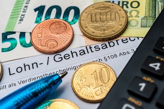 Gehalt Geldscheine- und Münzen liegen auf einem Zettel mit der Aufrschift "Lohn-/ Gehaltsabrechnung". Daneben liegen Kugelschreiber und Taschenrechner