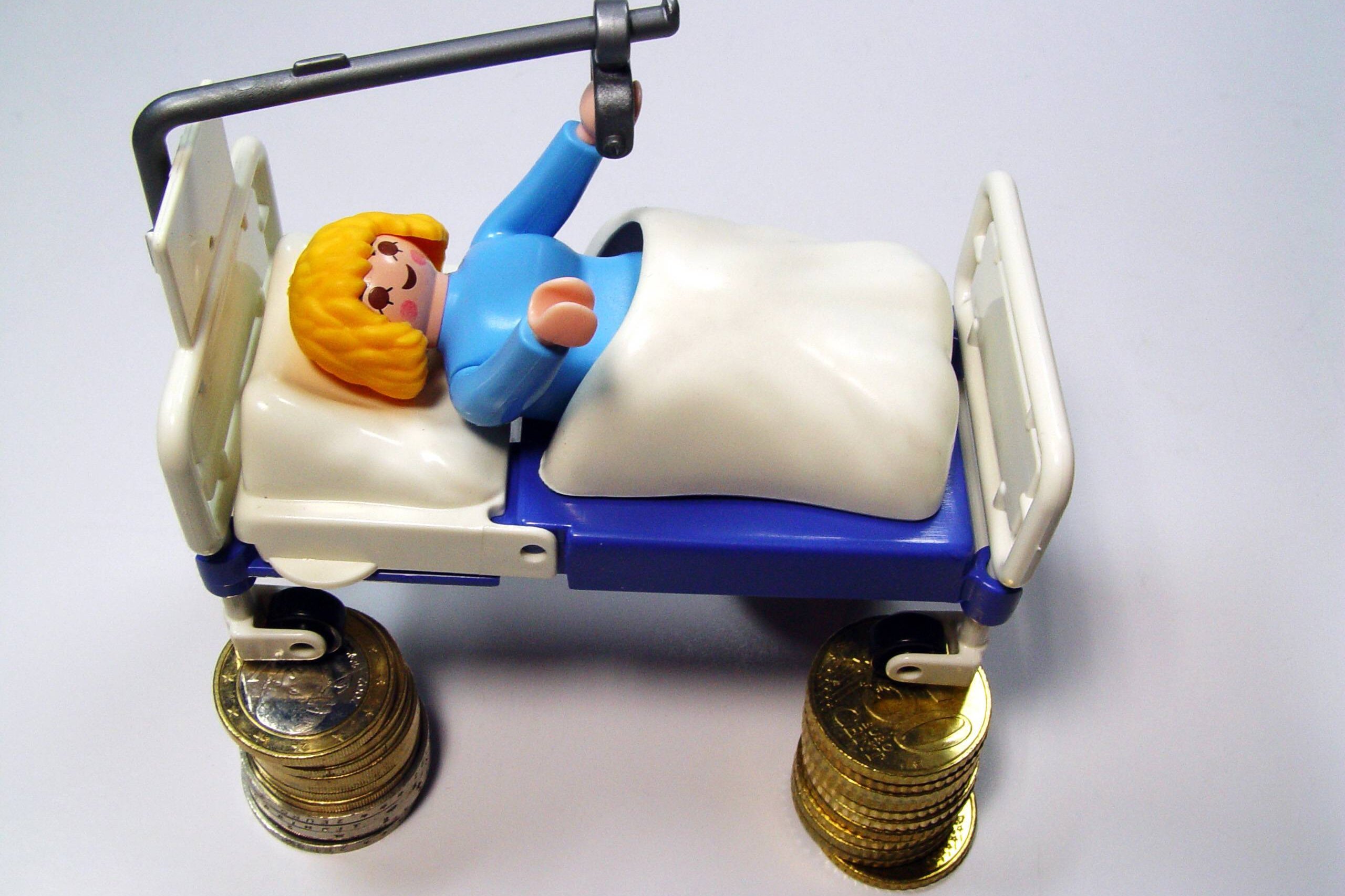 Ein Playmobil-Männchen in einem Krankenhausbett. Das Spielzeugbett steht auf kleinen Münzstapeln.