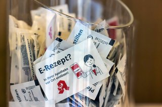 Ein großer Glasbehälter in einer Apotheke, gefüllt mit Werbegeschenken mit der Aufschrift "E-Rezept? Vor Ort in Ihrer Apotheke einlösen"