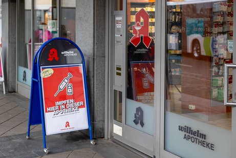 Impfung in der Apotheke Eine Apotheke mit einem Aufsteller vor dem Eingang, der auf die Möglichkeit einer Grippeschutzimpfung hinweist. Das Schild trägt die Aufschrift Impfen: Hier in dieser Apotheke und zeigt ein Symbol einer Spritze und eines Impfstoff-Fläschchens.