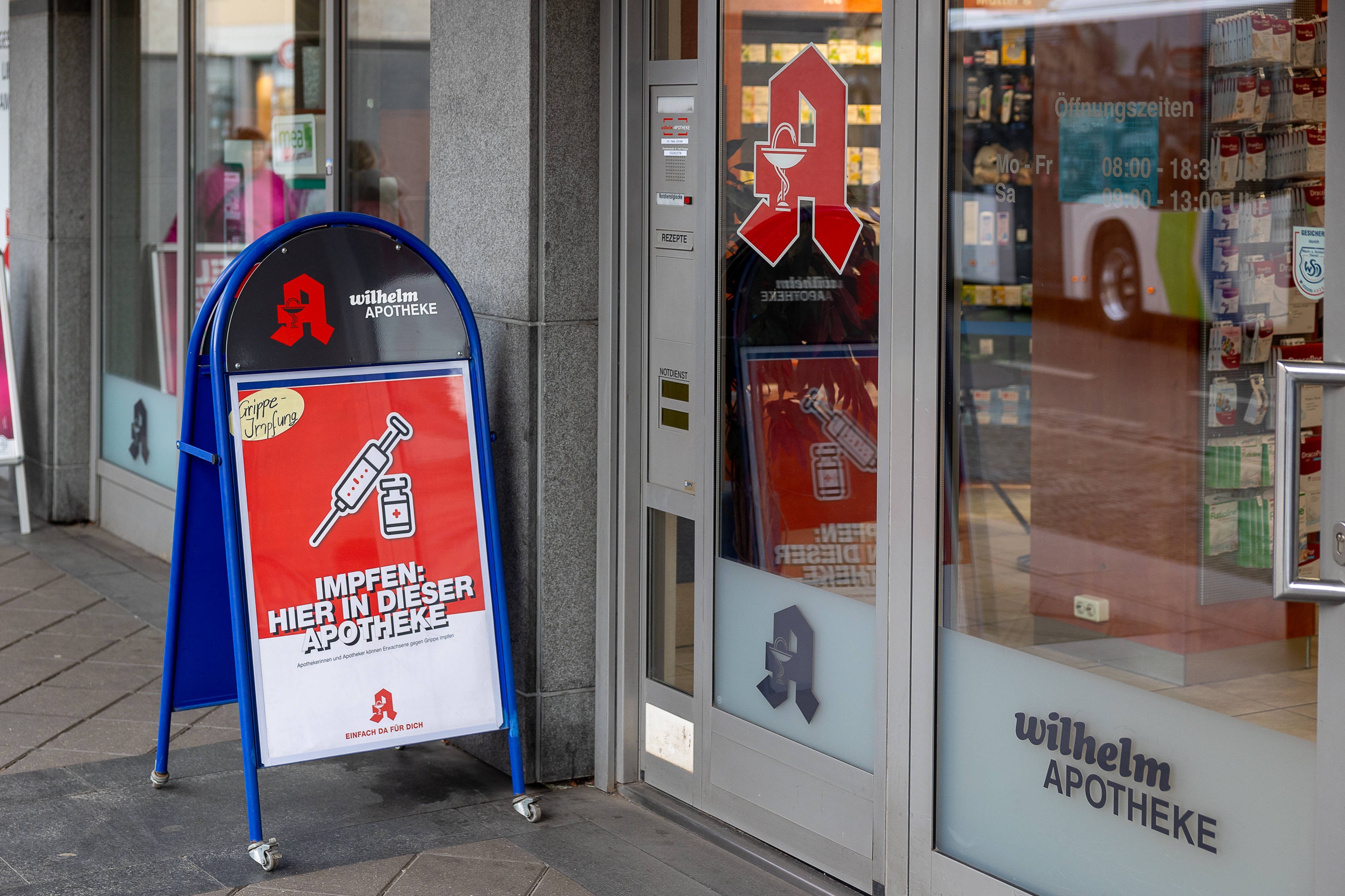 Impfung in der Apotheke Eine Apotheke mit einem Aufsteller vor dem Eingang, der auf die Möglichkeit einer Grippeschutzimpfung hinweist. Das Schild trägt die Aufschrift Impfen: Hier in dieser Apotheke und zeigt ein Symbol einer Spritze und eines Impfstoff-Fläschchens.