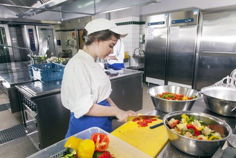 Eine junge Frau in Arbeitskleidung arbeitet in einer Großküche, sie schneidet Tomaten