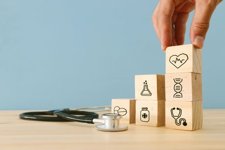 Eine Hand stapelt Würfel aufeinander, auf denen kleine Gesundheits-Icons zu sehen sind, z.B. ein Herz, ein Stethoskop und eine Arzneiflasche. Daneben liegt ein Stethoskop. 
