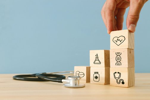 Eine Hand stapelt Würfel aufeinander, auf denen kleine Gesundheits-Icons zu sehen sind, z.B. ein Herz, ein Stethoskop und eine Arzneiflasche. Daneben liegt ein Stethoskop. 