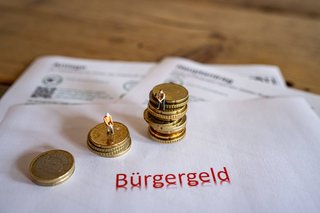 Ein Antrag für das Büregergeld, darauf unterschiedlich hohe Stapel von Münzen, auf denen kleine Figürchen sitzen. In roter Schrift steht darunter "Bürgergeld".