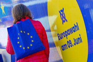 Eine Frau mit einer Tasche, die als Aufdruck die Flagge der Europäischen Union hat, steht vor einem blau-gelben Plakat mit der Aufschrift "Euriopawahl am 9. Juni". 