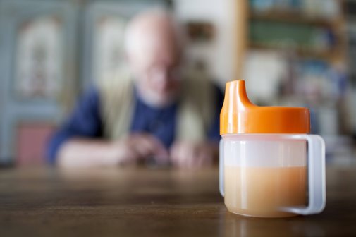 Pflege zu Hause Eine Schnabeltasse steht auf einem Tisch. Im Hintergrund sieht man unscharf einen alten Mann am Tisch sitzen.
