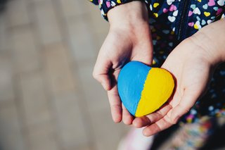 Hand eines Kindes hält einen herzförmigen Stein, der zur Hälfte blau und zur Hälfte gelb angemalt ist in den Farben der ukrainischen Flagge