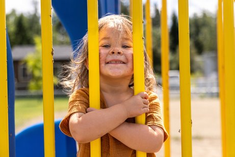 Ein kleines Mädchen auf einem Spielplatz, sie schaut durch Stäbe eines Spielgeräts, ihr Gesicht ist schmutzig