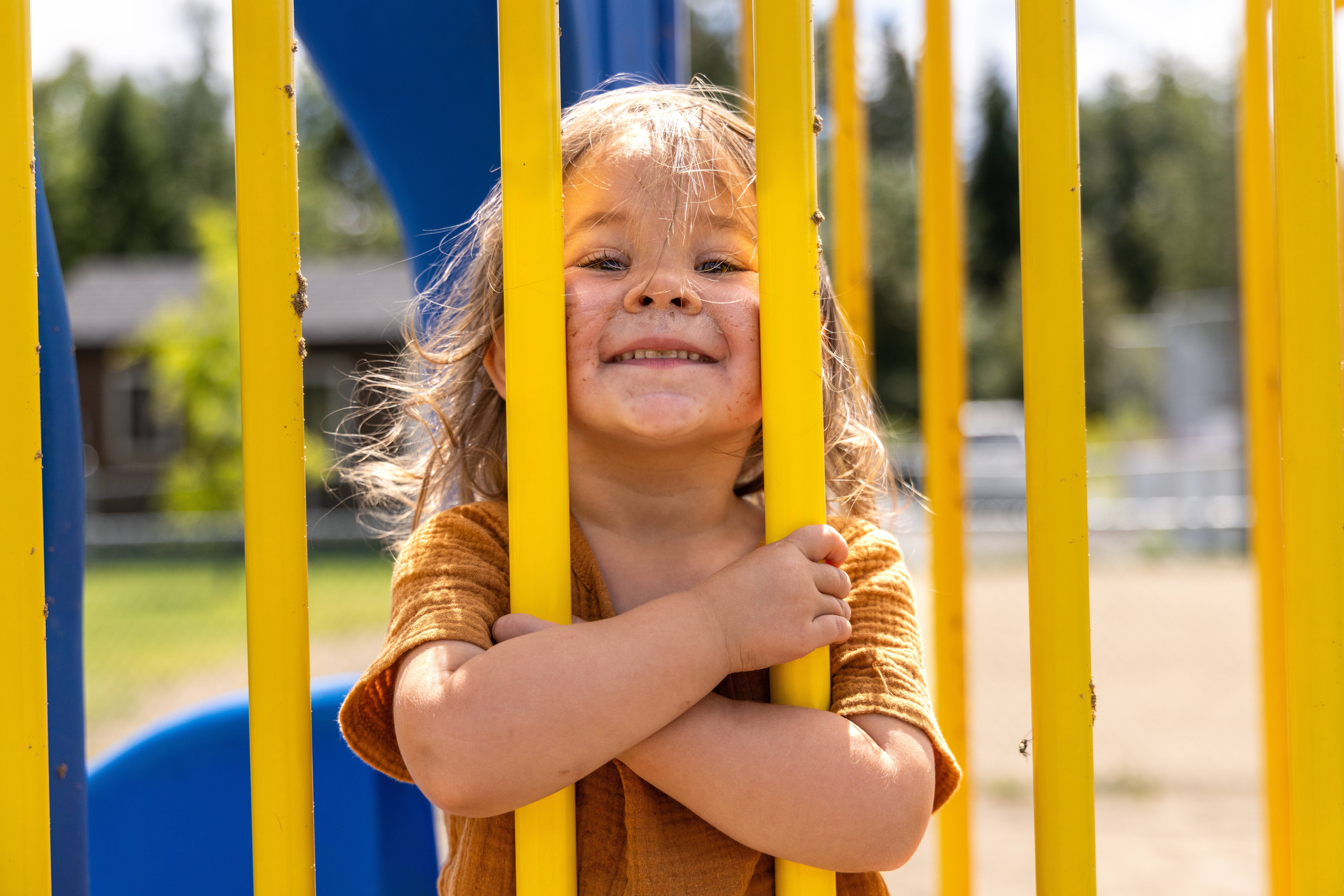 Ein kleines Mädchen auf einem Spielplatz, sie schaut durch Stäbe eines Spielgeräts, ihr Gesicht ist schmutzig