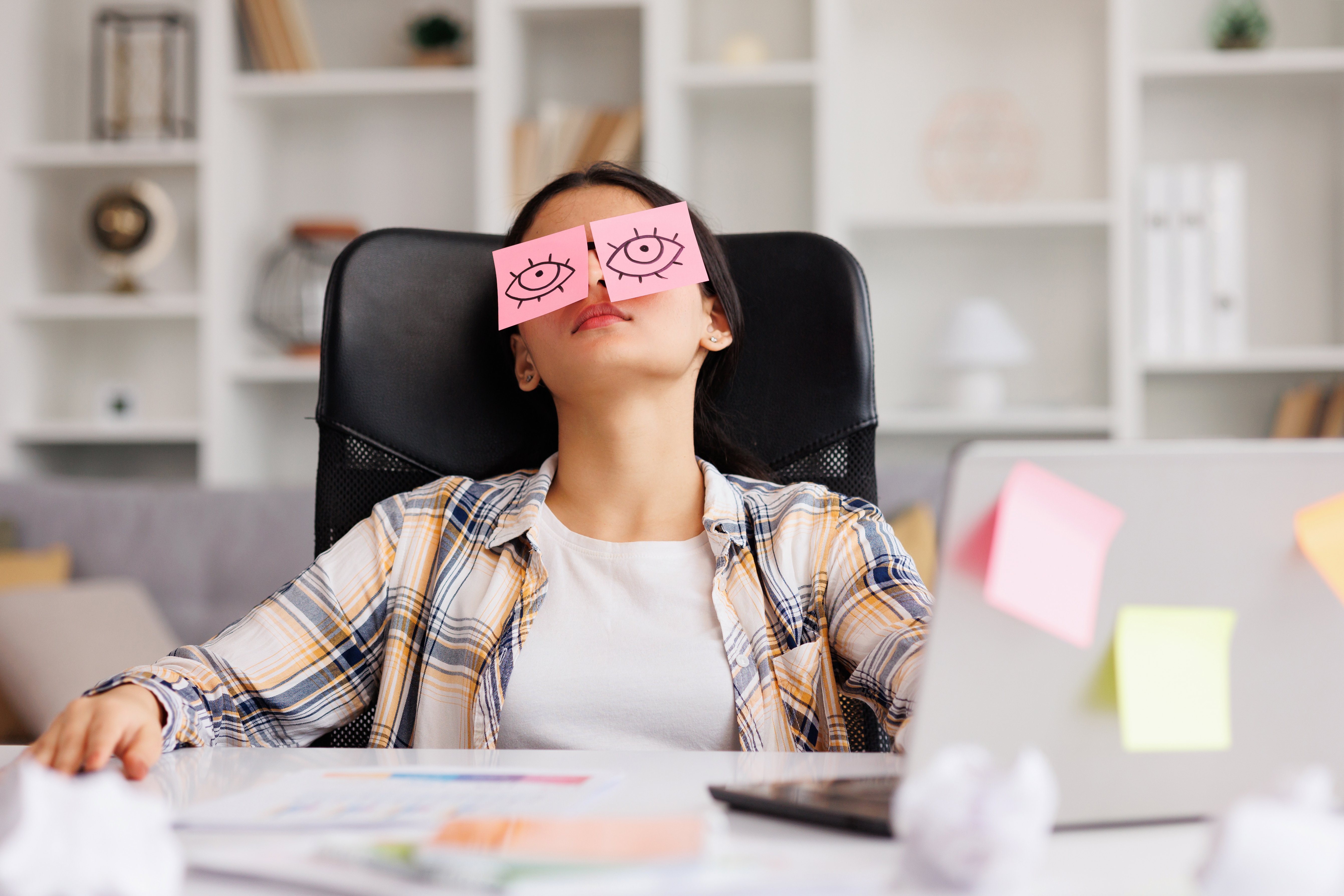 Belastung im Arbeitsleben Eine junge Frau sitzt am Schreibtisch in einem Büro, sie hat den Kopf zurückgelegt. Auf ihren geschlossenen Augen liegen Post-it-Zettel mit aufgemalten offenen Augen.