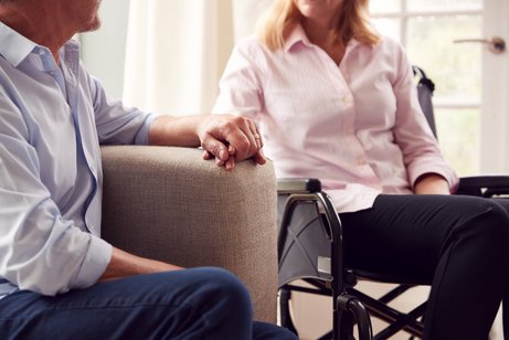 Mann und Frau halten sich an der Hand Älterer Mann auf einem Sofa und ältere Frau im Rollstuhl sitzen vertraut nebeneinander, halten sich an der Hand.
