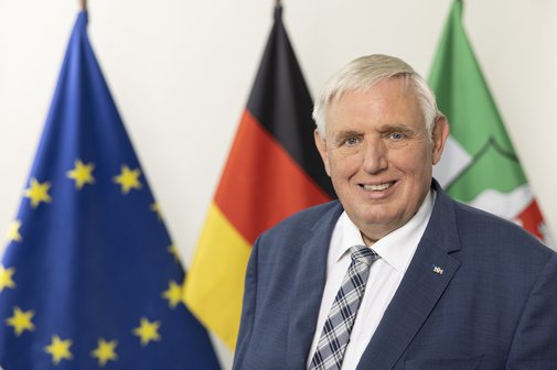 Das Foto zeigt Karl-Josef Laumann, im Hintergrund sind drei Flaggen zu sehen: Die Europa-Flagge, die Deutschlandflagge und die Flagge von Nordrhein-Westfalen