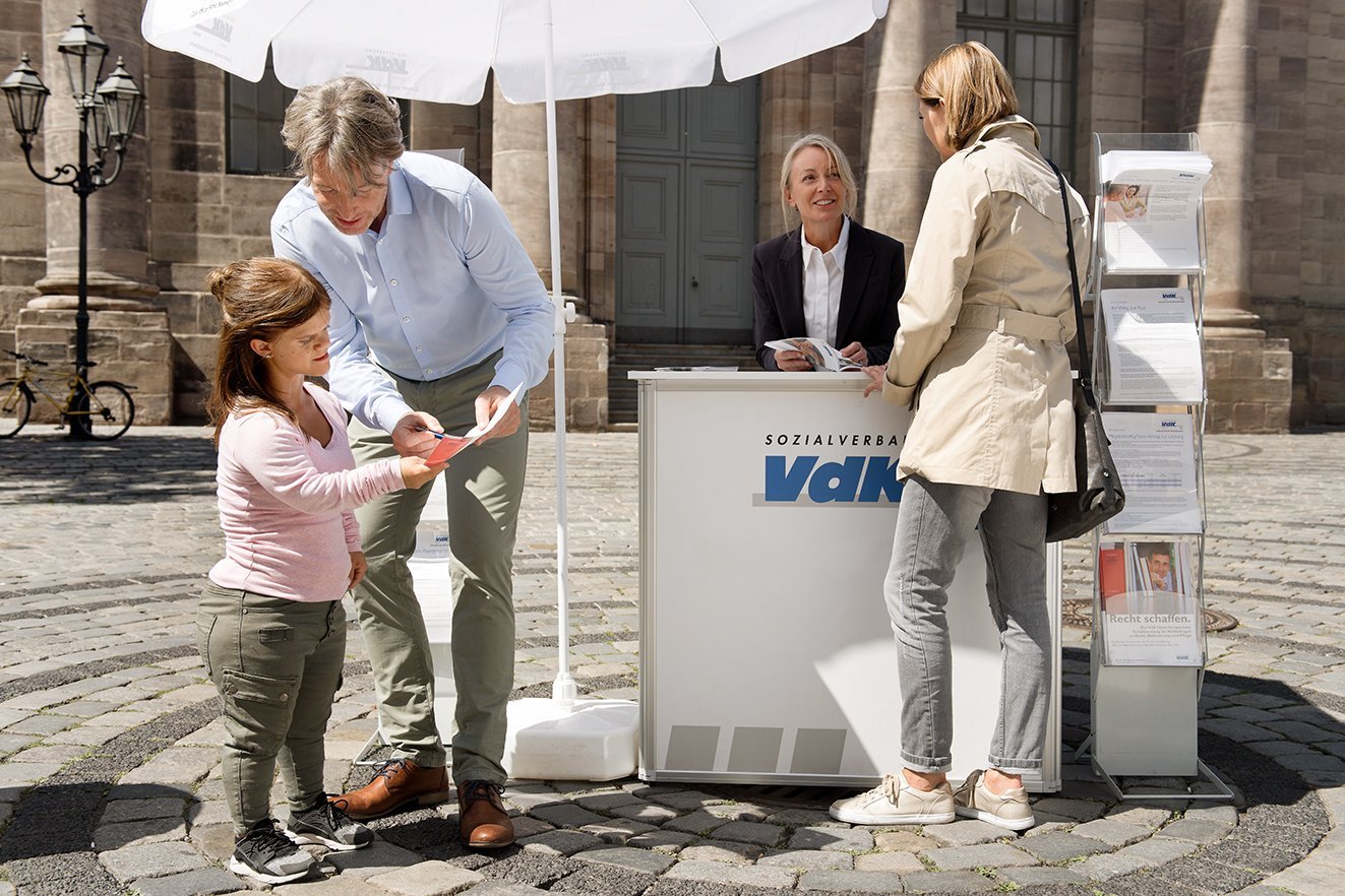VdK-Infostand Ein VdK-Infostand mit einer Mitarbeiterin, davor verschiedene Menschen, die sich informieren wollen.