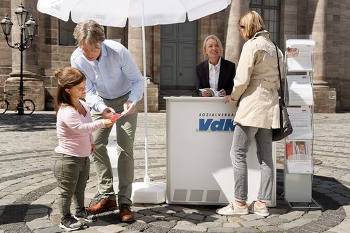 Ein VdK-Infostand mit einer Mitarbeiterin, davor verschiedene Menschen, die sich informieren wollen.