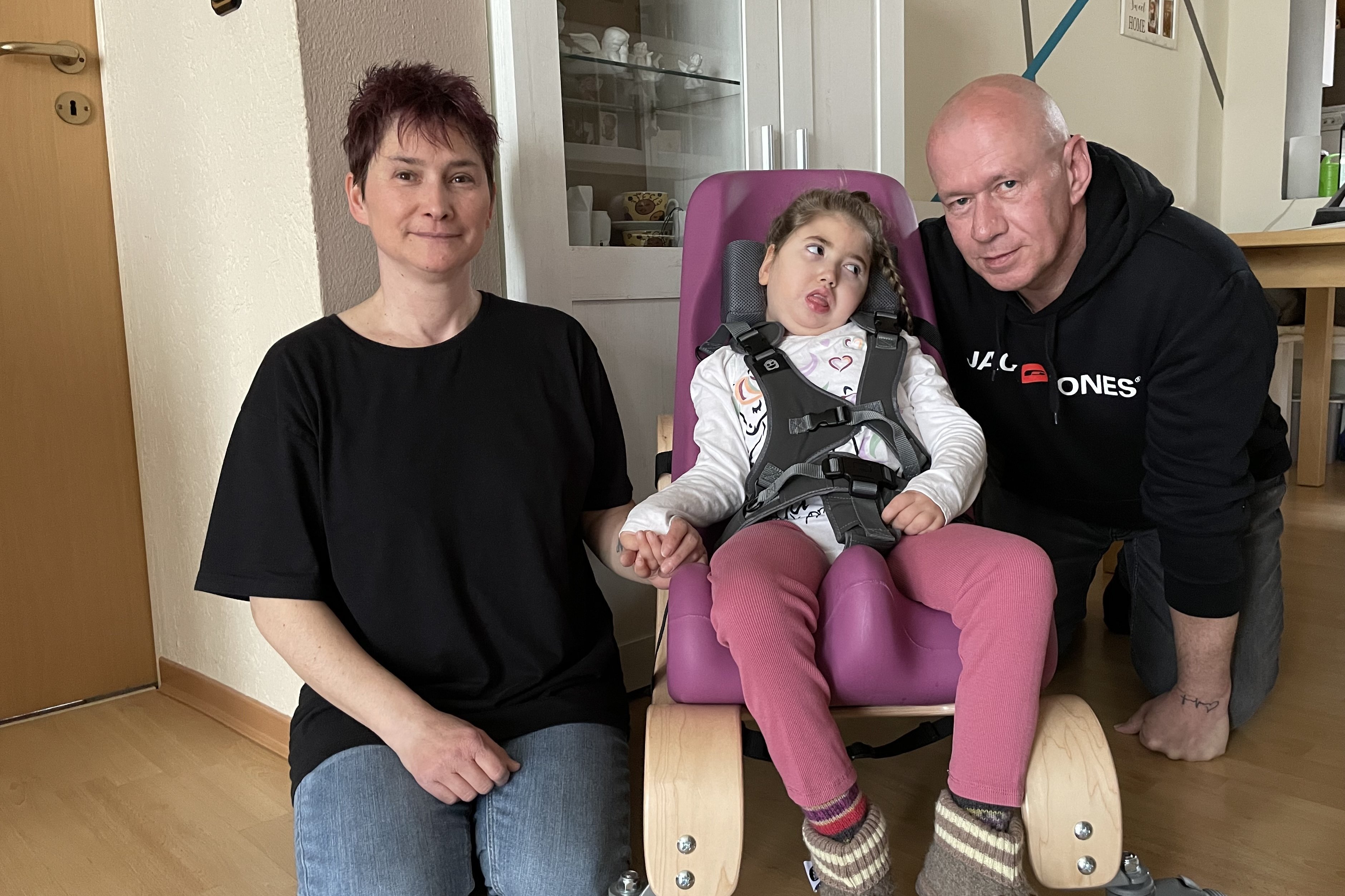 Das Ehepaar Heubner kniet links und rechts neben ihrer Tochter Nena am Boden, das Mädchen sitzt in dem speziellen Therapiestuhl, den der VdK durchsetzen konnte