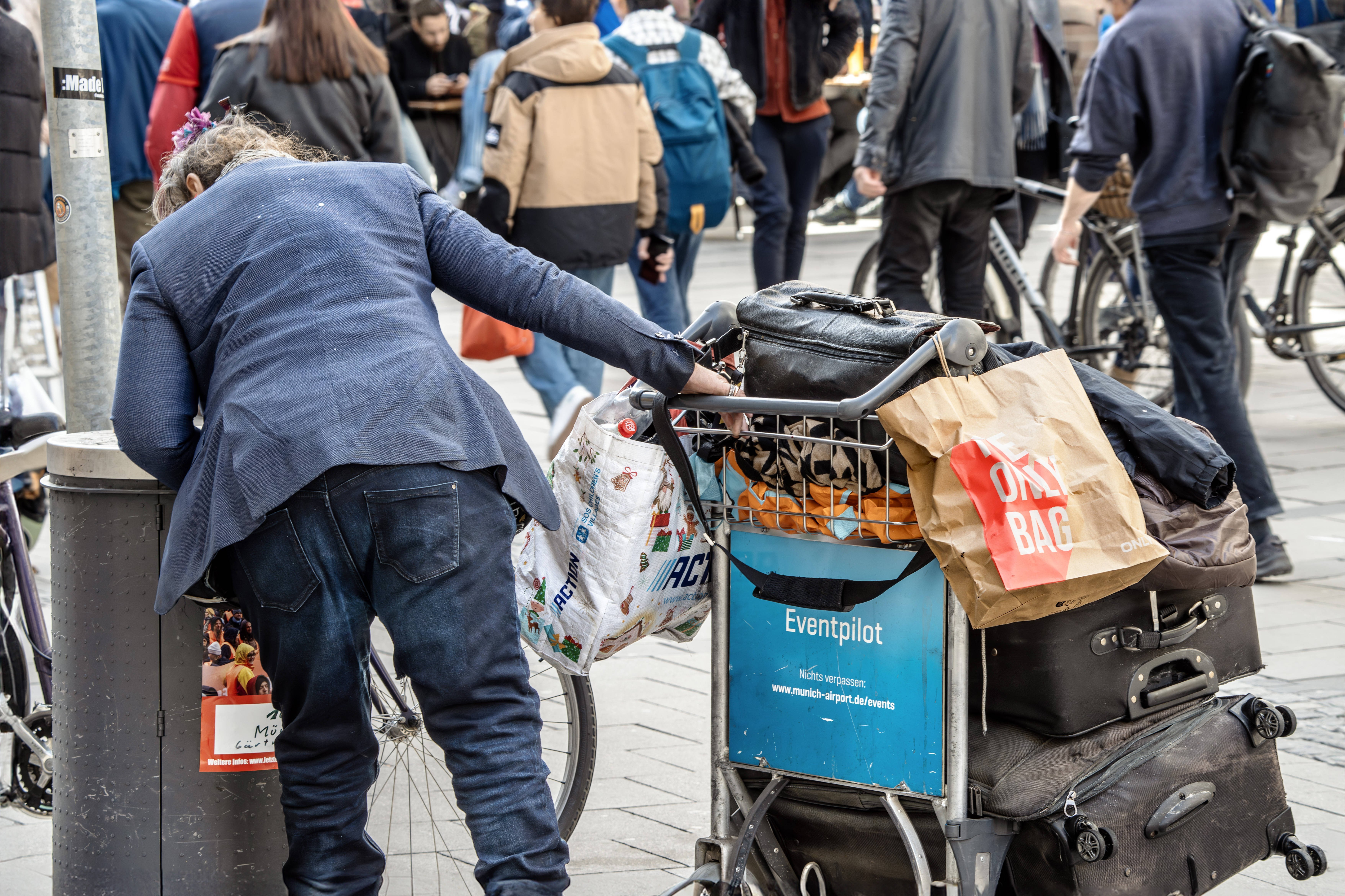 Flaschensammlerin Flaschensammlerin in der Innenstadt von München - eine Frau mit einem Einkaufswagen voller gesammelter Flaschen und anderer Dinge beugt sich über einen Mülleimer, schaut hinein.