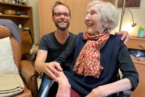 Dirk Angele und Anita Göbels, die Bewohnerin des Seniorenheims, in dem Dirk Angele ehrenamtlich tätig ist. Sie sitzt im Rollstuhl, er sitzt hinter ihr, legt eine Hand auf ihre Schulter und die andere auf ihren Arm.