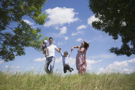 Eine junge Familie mit zwei kleineren Kindern auf einer Wiese vor blauem Himmel, der Vater tr&auml;gt ein Kind auf dem Arm, das andere Kind halten die Eltern zwischen sich und lassen es "fliegen".