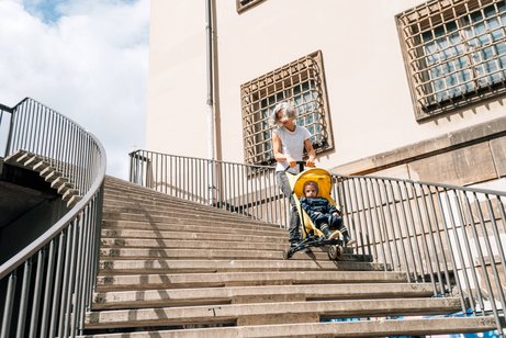 Frau mit Kinderwagen bei Hitze Eine Frau fährt mühevoll mit einem Buggy mit Kleinkind treppab, die Sonne scheint, sie wirkt erhitzt und angestrengt