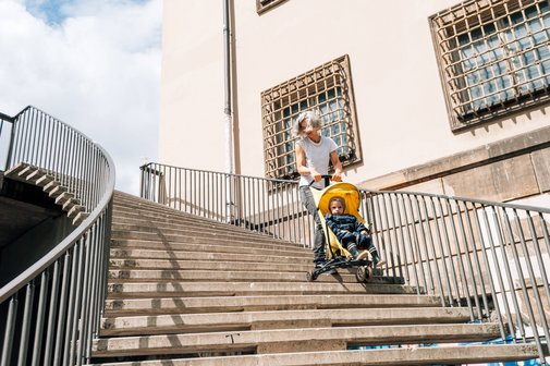 Eine Frau fährt mühevoll mit einem Buggy mit Kleinkind treppab, die Sonne scheint, sie wirkt erhitzt und angestrengt