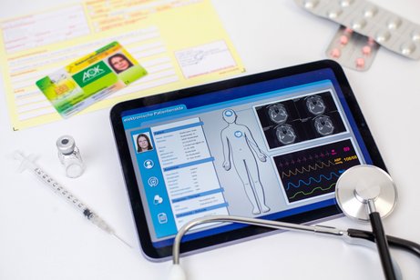 Elektronische Patientenakte ePA Ein Tablet liegt auf einem Tisch, darauf sind medizinische Patientendaten zu sehen. Daneben liegen ein Stethoskop, eine Versichertenkarte, Blister mit Tabletten und Arztdokumente und Rezepte.