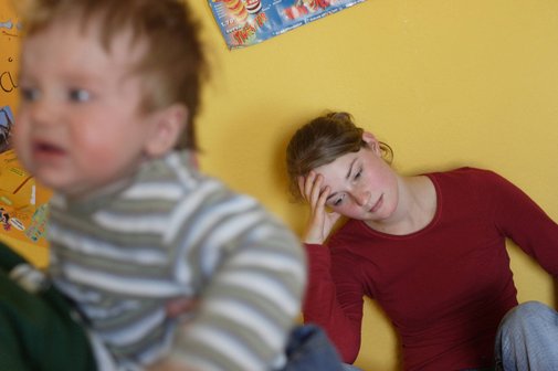Alleinerziehende Mutter Ein Kleinkind im Vordergrund, im Hintergrund ein junge Frau, die am Boden sitzt und den Kopf erschöpft in die Hand gestützt hat
