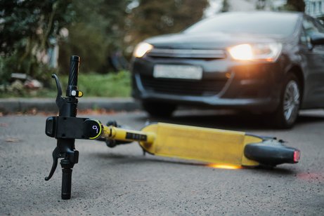 Haftung Unfall E-Scooter Ein gelber E-Scooter liegt umgestürtzt auf der Straße. Ein Auto, dessen Scheinwerfer an sind, steht weniger Meter entfernt.