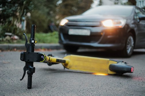Haftung Unfall E-Scooter Ein gelber E-Scooter liegt umgestürtzt auf der Straße. Ein Auto, dessen Scheinwerfer an sind, steht weniger Meter entfernt.