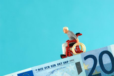 Pflege und Armut Symbolbild: Ein 20-Euro-Schein bildet eine abschüssige Rampe, auf der eine Mini-Figur eines alten Mannes im Rollstuhl bergab rollt