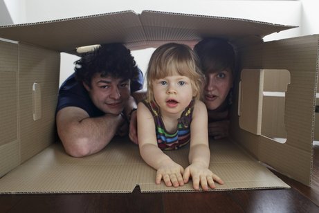 Eine junge Familie: Vater, Mutter und ein kleines Mädchen schauen gemeinsam aus einem großen Pappkarton heraus, der auf dem Boden liegt.