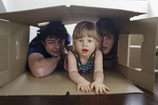 Eine junge Familie: Vater, Mutter und ein kleines Mädchen schauen gemeinsam aus einem großen Pappkarton heraus, der auf dem Boden liegt.